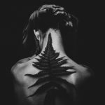 Black and white photo of a tattooed woman from the back, featuring a fern design. Captivating and artistic pose.